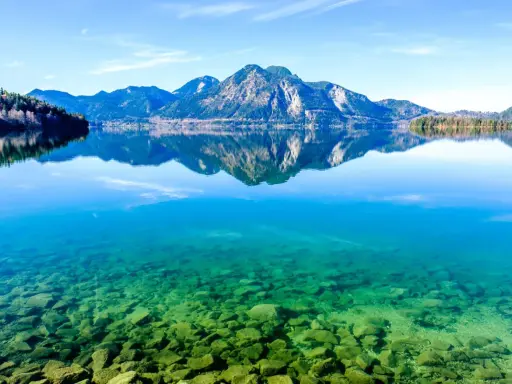 Kristallklarer Badesee in Bayern mit Felsen am Ufer und spiegelnden Bergen im Hintergrund.