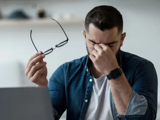 Homme fatigué, lunettes à la main, se frottant les yeux devant un ordinateur. Il est temps de penser à comment protéger ses yeux des écrans.