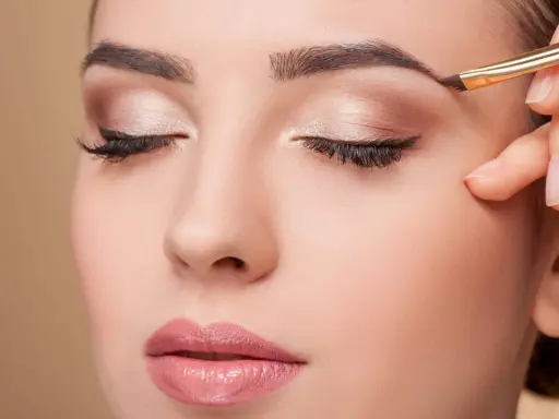 Une main applique un maquillage subtil pour parfaire la forme sourcil d'une femme aux yeux fermés.
