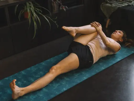 Femme effectuant un étirement psoas sur un tapis de yoga bleu. Elle est allongée sur le dos, une jambe pliée et ramenée vers la poitrine.
