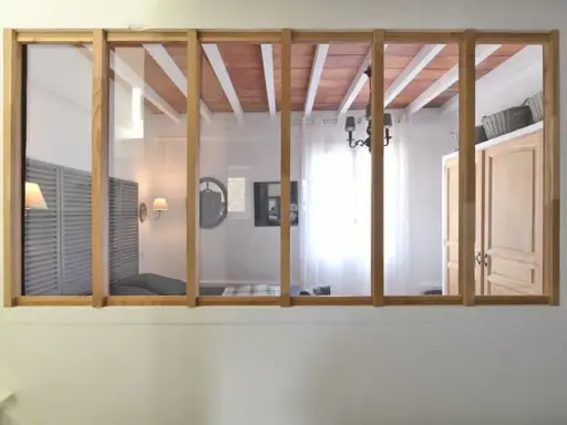 Une verrière en bois sépare deux pièces. On y voit un lit, des volets, une armoire et un lustre.