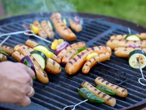 Grilla korv med grönsaker på en kolgrill. Röken stiger från den saftiga maten.