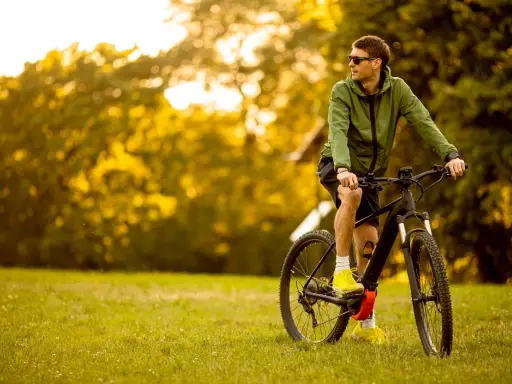 Mężczyzna w zielonej kurtce i okularach przeciwsłonecznych odpoczywa na trawie obok swojego roweru elektrycznego ebike.