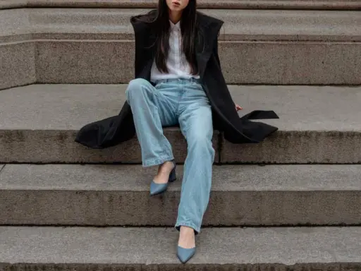 Donna con cappotto nero, camicia bianca e **pantaloni a sigaretta** chiari, indossa **tronchetti** blu.