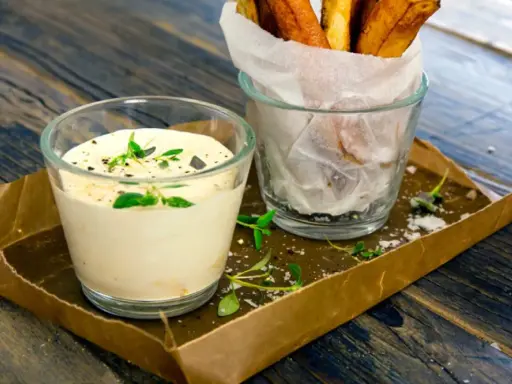 En krämig creme fraiche sås med örter serveras bredvid en glasbehållare med krispiga pommes frites på ett rustikt träbord.
