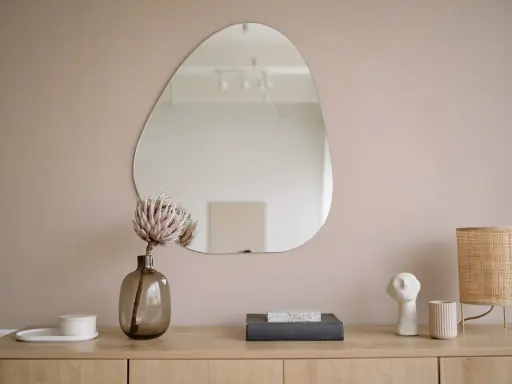 Un miroir vitrail aux formes organiques reflète une pièce épurée. Une fleur séchée dans un vase en verre fumé et une lampe en rotin complètent la scène.