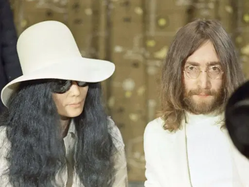 John Lennon och Yoko Ono, ikoniska konstnärer, poserar tillsammans. Yoko bär en stor vit hatt och solglasögon, medan John har sitt karaktäristiska långa hår och glasögon.