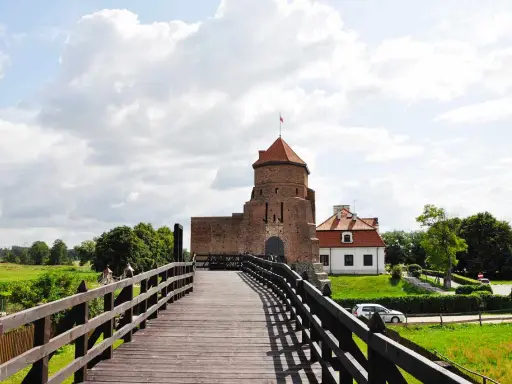 Drewniany most prowadzi do ceglanego zamku z czerwoną wieżą i białym budynkiem.