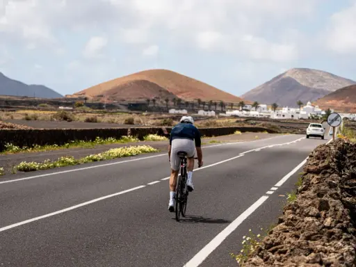 Ein Radfahrer auf einem Rennrad fährt auf Lanzarote durch eine vulkanische Landschaft mit weißen Häusern und Palmen.
