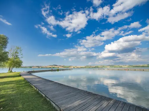 Passeggiata in legno sul **lago di Chiusi**, con cielo azzurro e nuvole bianche che si riflettono sull'acqua calma.