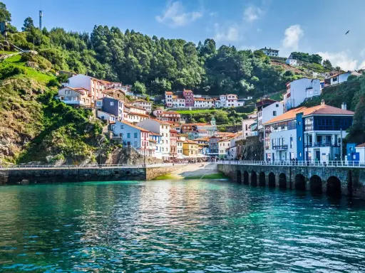 Un pintoresco pueblo costero con **5 villas** coloridas apiladas en una colina verde, frente a un mar turquesa.