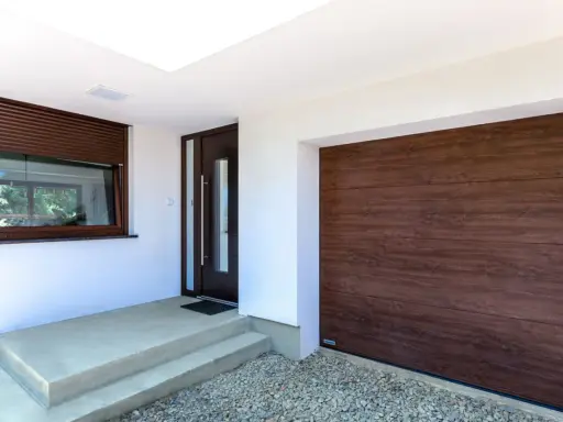 Ingresso con porta scura e avvolgibili marroni, accanto a un'ampia porta garage in legno scuro.