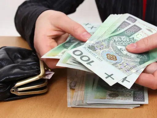 Dłonie liczą banknoty 100 zł obok portmonetki. Zastanawiasz się, ile wynosi trzynasta emerytura netto?