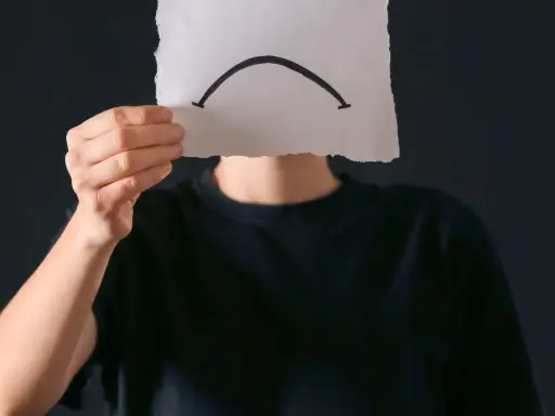 Osoba w czarnej bluzie trzyma kartkę z narysowaną smutną buźką. Czy to anhedonia, co to za uczucie?