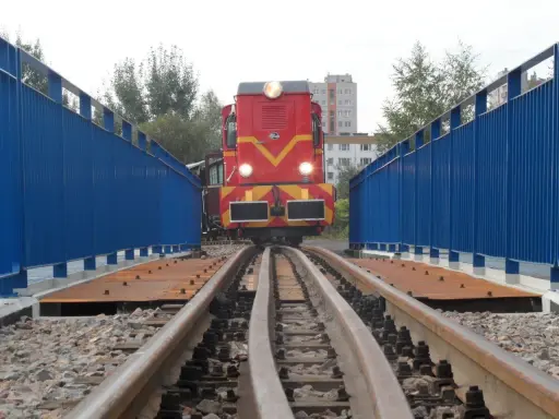 Czerwona lokomotywa na torach wąskotorowych, część górnośląskich kolei wąskotorowych, wjeżdża na most z niebieskimi barierkami.