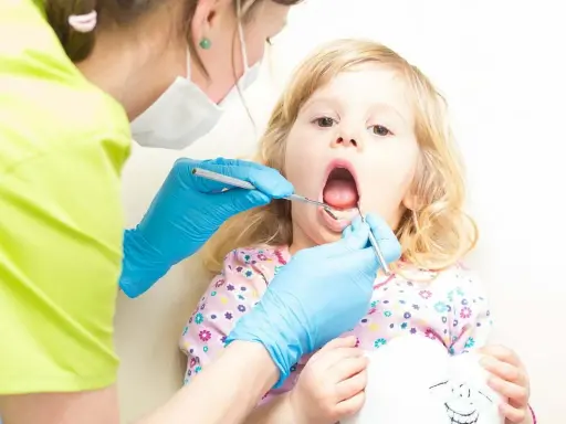 Mała dziewczynka z otwartymi ustami, kiedy z dzieckiem do dentysty. Stomatolog w niebieskich rękawiczkach bada jej ząbki.