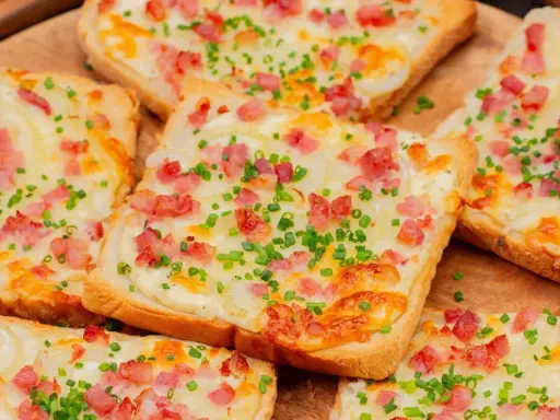 Mehrere Flammkuchen-Toast-Scheiben mit Speck, Zwiebeln und Schnittlauch belegt, auf einem Holzbrett.