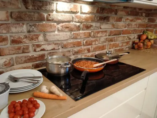 Cegła w kuchni inspiracje: gotowanie na płycie indukcyjnej, obok garnki, patelnia z sosem, warzywa i owoce.