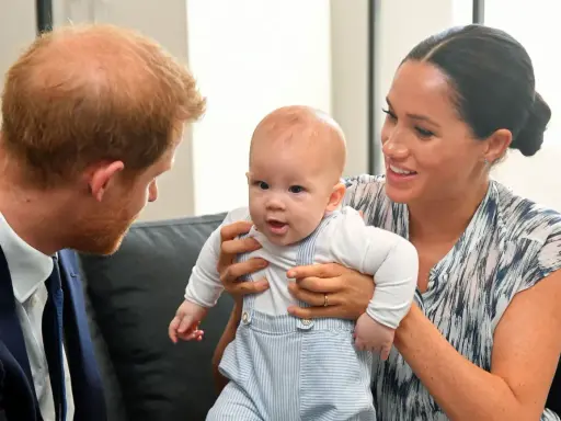 Meghan Markle håller sitt barn, som tittar nyfiket framåt. Prins Harry ser på dem med ett leende.
