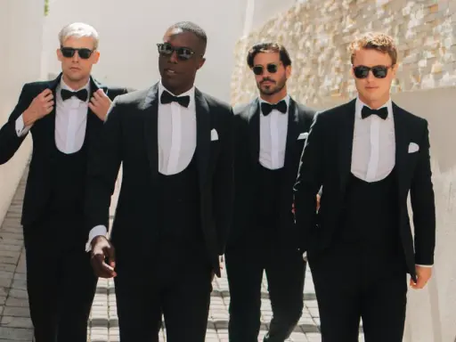 Four men stride confidently in a modern black tie dress code, sporting sharp tuxedos and sunglasses.