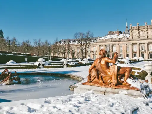 Schneebedeckte Gärten und ein gefrorener Brunnen vor einem Palast in Spanien im Winter. Eine bronzene Statue blickt auf die verschneite Landschaft.