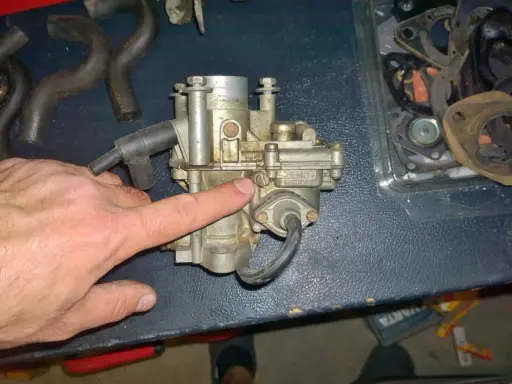 Main d'homme pointant un carburateur Zenith, possible cause d'un moteur 4 temps qui s'étouffe à l'accélération. Pièces de rechange visibles.