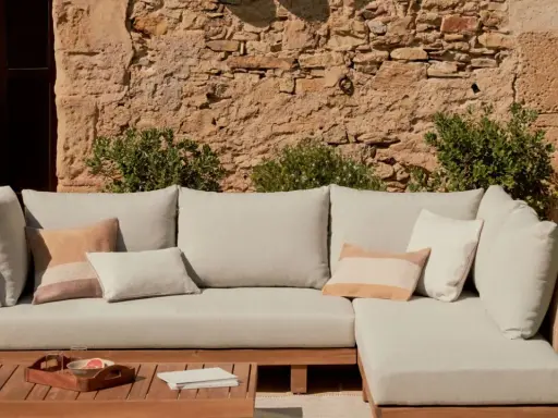 Sofá modular de exterior con cojines en tonos neutros, sobre una mesa de madera. La **decoración estilo natural** se realza con la pared de piedra y la vegetación.