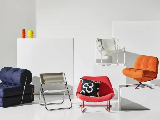 En samling möbler i 70-tals inredning: en blå bäddsoffa, en röd fåtölj med mönstrat kudde, en orange fåtölj och en vit metallstol.