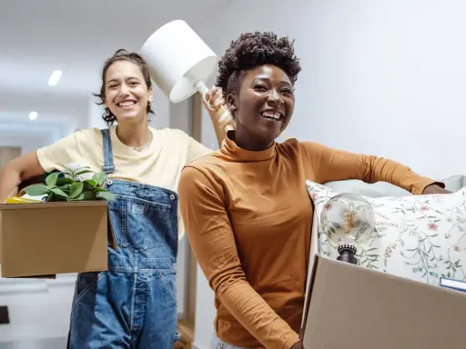 Deux amies souriantes déménagent, portant des cartons remplis d'objets. Une nouvelle aventure commence, peut-être grâce à un **bail solidaire**.
