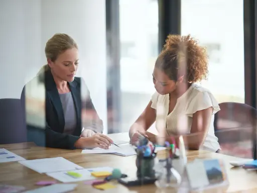 Deux femmes discutent de documents lors d'une réunion. L'une, blonde, porte un blazer sombre. L'autre, aux cheveux bouclés, est en chemisier clair. Leurs mains pointent vers les papiers, comme pour en comprendre le **signification**.