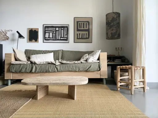 Une idée table basse fait maison en bois brut, posée sur un tapis tressé, devant un canapé vert et des tableaux abstraits.
