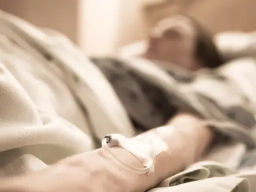 Ręka pacjenta z wenflonem w szpitalnym łóżku, w tle niewyraźna postać.