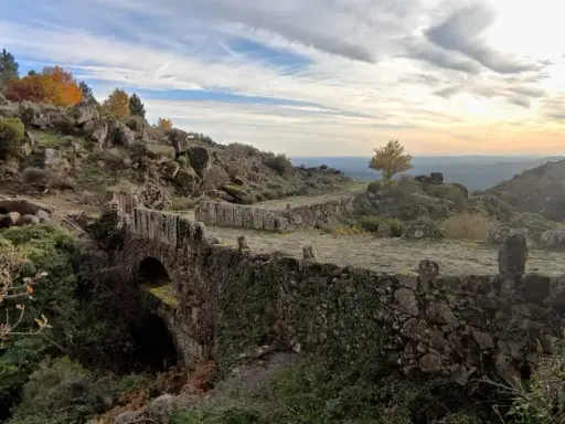 Herbstliche Farben in der Sierra de Gata. Ein alter Steinbogenbrücke, überwuchert von Efeu, führt durch eine felsige Landschaft.