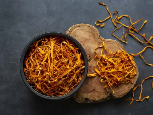 Un bol de cordyceps séchés, connus pour leurs nombreux bienfaits, est posé sur un fond sombre. Quelques brins dorés s'échappent du bol.