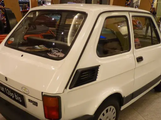 Biały Fiat 126p bis z naklejkami na tylnej szybie, stojący obok czerwonego samochodu.
