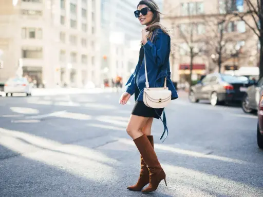 A woman walks down a city street wearing a black dress, a denim jacket, and stylish brown boots.