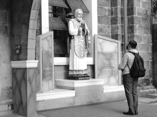 Pomnik Ojca Pio, patrona modlących się o uzdrowienie, stoi przed kościołem. Mężczyzna z plecakiem patrzy na tablicę.