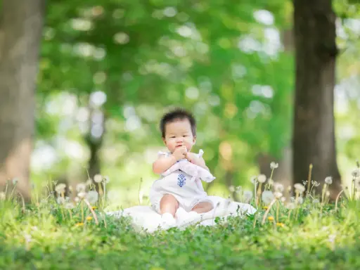 Dolce bimbo di 9 mesi seduto sull'erba, con soffioni e alberi sullo sfondo. Una foto che cattura la sua curiosità.