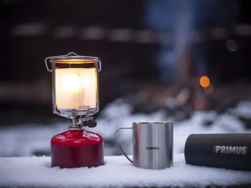 En varm primus lampa lyser upp snön bredvid en mugg och en termos. I bakgrunden syns en lägereld.