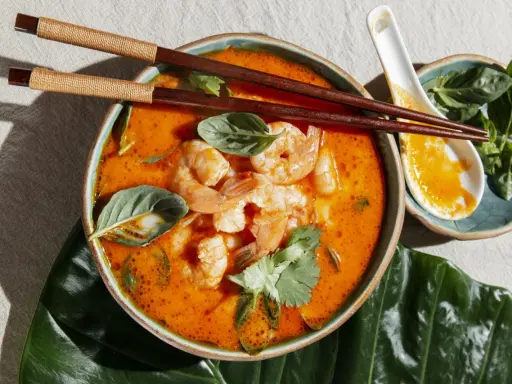 Miseczka zupy tom yum z krewetkami, ozdobiona ziołami, pałeczki i łyżka.