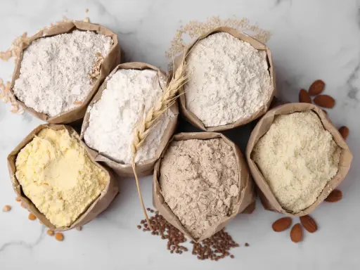 Six sacs en papier remplis de différentes farines, dont une farine de blé CRC. Amandes et grains parsèment le fond marbré.