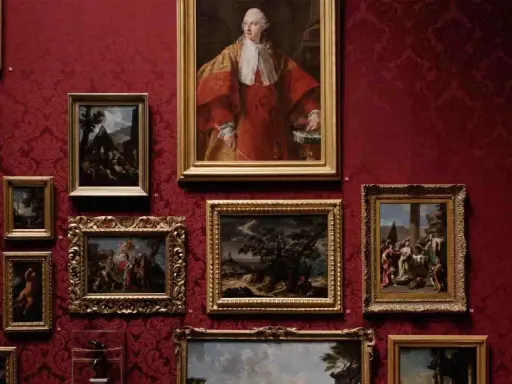 A gallery wall displays classical paintings in ornate frames against a rich red damask background. This collection showcases the best way to invest in art, with portraits and landscapes hinting at historical narratives.