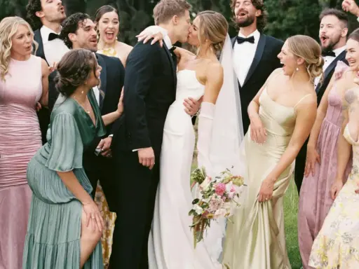 A wedding party celebrates as the bride and groom kiss. The groomsmen attire is classic black tuxedos.