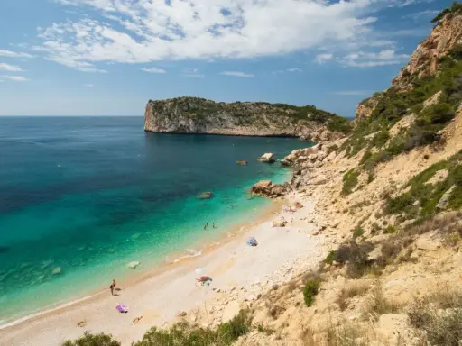 Türkisblaues Wasser trifft auf Kiesstrand in einer malerischen spanischen Atlantikbucht. Felsen und grüne Vegetation säumen die Küste.
