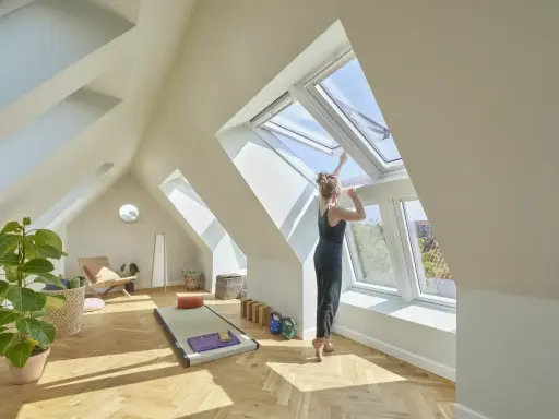 Frau öffnet Dachfenster in hellem Dachboden-Yoga-Raum. Hier darf man Dachboden als Wohnraum nutzen und schafft sich eine Oase.