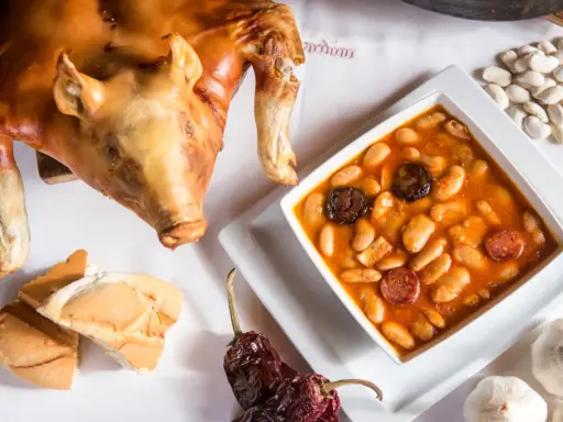 Cochinillo asado segoviano, crujiente y dorado, acompañado de un plato de judiones con chorizo y morcilla, pan y ajos.