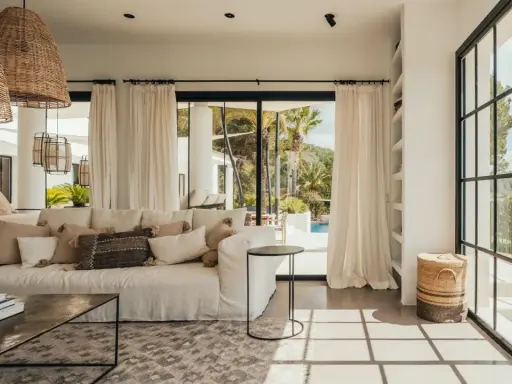 Arredare una casa al mare: divano bianco, cuscini naturali, lampade in rattan e vista su piscina e palme.