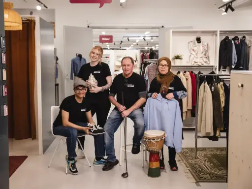 Fyra personer poserar i en butik med kläder och föremål. En kvinna håller en vas, en man en golfklubba, och en annan kvinna en ljusblå hoodie. De representerar hållbart second hand.