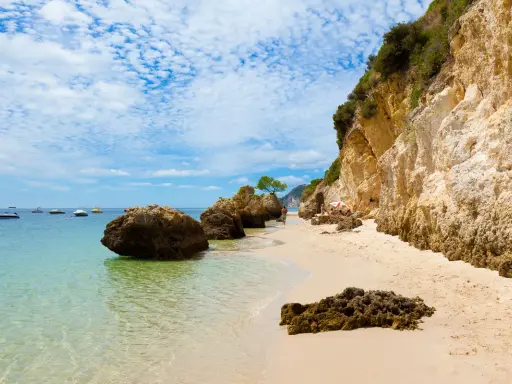 Sonniger Setúbal Strand mit türkisfarbenem Wasser, Felsen und Booten im Hintergrund. Eine kleine Gruppe entspannt sich unter einem Sonnenschirm.