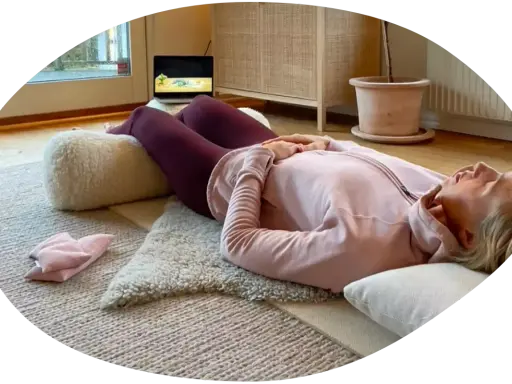 Kvinna ligger på rygg på en matta med en kudde under knäna, hon försöker varva ner med en laptop i bakgrunden.